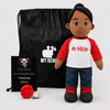 A plush doll with dark skin, black hair, a red-and-white shirt reading 'My Hero,' blue jeans, and black shoes stands in front of a black drawstring bag and a small black card on a light gray background.