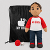 A plush doll dressed in a red and white raglan shirt with 'My Hero' printed in red, blue jeans, and dark shoes, standing beside a black drawstring bag with a white soldier graphic and 'MY HERO' text, plus a small red helmet and white cord on a light gray background.