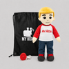 A plush doll with blond hair, a red and white shirt with 'MY HERO' text, blue jeans, and black shoes, standing next to a black drawstring bag and a small red accessory on a light gray background.