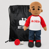 A plush doll toy with a bald head and brown skin, wearing a red and white raglan shirt that says 'MY HERO' in red, blue jeans, and black-and-red shoes, shown with a black drawstring bag and a red circular accessory on a light gray background.