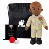 A firefighter-themed plush doll stands next to a black drawstring bag, a black gift card, and a red fire alarm on a white background. The doll wears a tan firefighter uniform with yellow stripes and black boots.