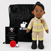 A firefighter-themed plush doll stands in front of a black drawstring bag and a black thank-you card. The doll is wearing a tan uniform with yellow reflective stripes and black boots.