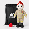 A firefighter-themed plush doll wearing a tan uniform, red beret, and black boots, standing in front of a black drawstring bag with a white firefighter silhouette and the words 'MY HERO'. A red helmet and a white USB charging cable are placed in front on a light gray background.