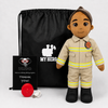 A firefighter-themed plush doll stands in front of a black drawstring bag, a black card reading 'MY HERO,' and a red fire alarm on a light gray studio background. The doll wears a tan firefighter uniform with yellow reflective stripes and black boots.