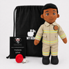 A firefighter-themed plush doll stands in front of a black drawstring bag, with a black gift card and a red fire helmet accessory on a light gray studio background. The doll wears a tan uniform with yellow reflective stripes and black boots.