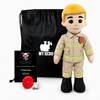 A firefighter-themed plush doll stands in front of a black drawstring bag, with a small black thank-you card and a red fire hydrant accessory on a white studio background. The doll wears a tan uniform with yellow reflective stripes, black boots, and a yellow hair color.