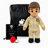 A plush firefighter doll stands in front of a black drawstring bag, with a small black card and a red fire helmet on a white studio background. The doll wears a tan firefighter uniform with yellow reflective stripes, black boots, and a brown cap.