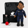 A Police doll dressed as a police uniform stands next to a black drawstring bag, a black card reading 'UMBO. From our military family to yours, THANK YOU!', and a small red ribbon on a light gray background.