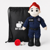 A plush toy dressed as a police officer stands next to a black drawstring bag with a white firefighter graphic and text, with a red helmet and white USB charging cable in front, on a light gray background.