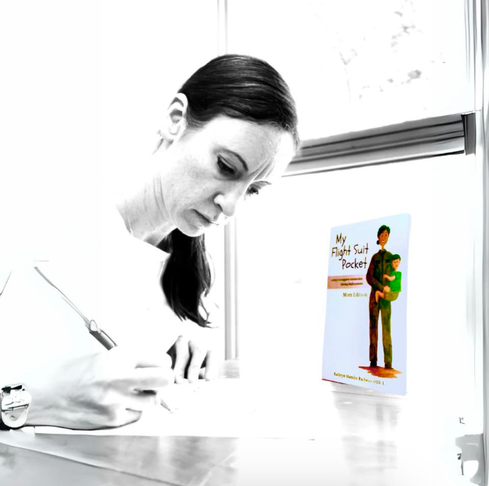 Kathryn “Katie” Hamlin-Pacheco is writing at a desk, with a copy of the book 'My Flight Suit Pocket' standing open on the table beside her. The photo has a selective-color effect, rendering the woman and desk in black and white while the book cover retains its natural color.