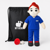 Nurse doll in blue medical scrubs with fair skin and red hair, female recordable medical plush doll. A plush doctor doll wearing blue scrubs and a red cap stands in front of a black drawstring bag with a white 'MY HERO' graphic. A red circular accessory and a small white USB charging cable sit on the floor in front, all on a light gray background.
