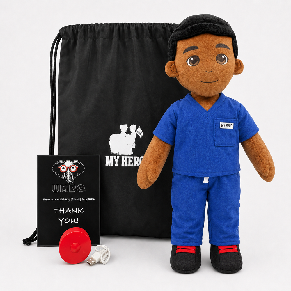 A plush doctor doll stands in front of a black drawstring bag and a small black gift card. The doll has brown skin, dark hair, a black cap, a blue scrub top, matching blue pants, black shoes with red soles, and a small white chest patch reading 'MY HERO'.